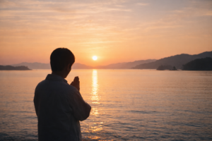 朝日が昇る海に向かって手を合わせ祈る人のシルエット